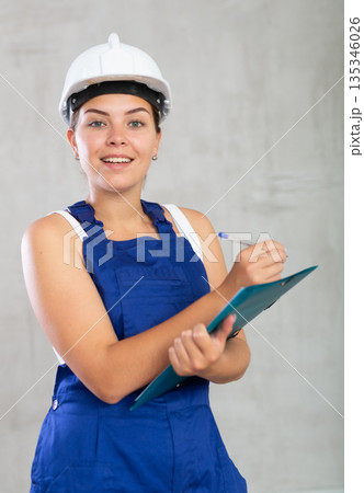 Portrait of emotional mechanic girl in overalls and a protective helmet, who writes in documents Portrait of emotional mechanic girl in overalls and a protective helmet, who writes in documents 135346026