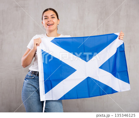 Joyous young woman holding Scotland flag against unicoloured background 135346125