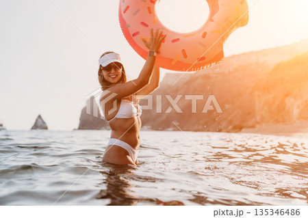 Woman inflatable beach. Young woman enjoys summer vacation holding a donut shaped ring in the sea near cliffs. 135346486