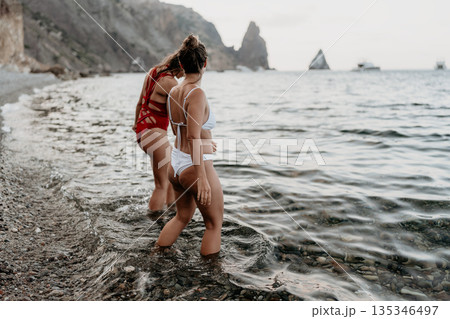 Women, beach, sea. Two women wade in shallow water on a rocky beach by majestic cliffs, enjoying a serene summer. 135346497