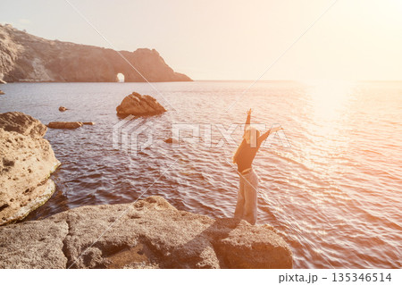 Woman sunset sea standing on a rocky cliff with arms raised towards the sun enjoying a moment of freedom and serenity 135346514