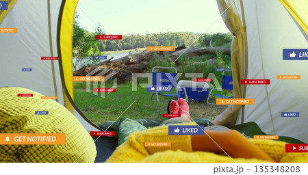 Relaxing camper lying in sleeping bag in yellow tent by lakeside clearing, with chairs and cooler 135348208