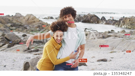 Smiling couple hugging on rocky shoreline in jeans and yellow sweater, with pebbles and ocean 135348247