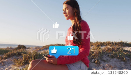 Sitting woman in sweater and shorts holding smartphone on dune viewing thumbs up icons, copy space 135348260