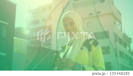 Mature Middle Eastern woman wearing hijab holding smartphone in outdoor plaza with green overlay Mature Middle Eastern woman wearing hijab holding smartphone in outdoor plaza with green overlay 135348295