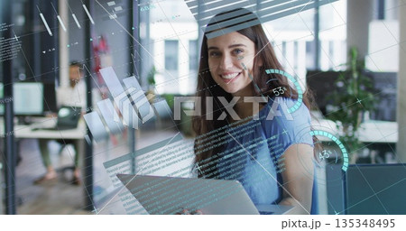 Working woman sitting at desk smiling in open-plan office, with laptop, holographic data overlays 135348495
