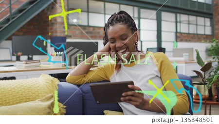 Smiling woman wearing yellow shirt holding tablet on blue sofa in office, with FLASH SALE overlays 135348501