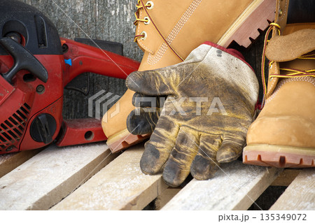 Chainsaw, work gloves, and sturdy boots resting on a wooden pallet Chainsaw, work gloves, and sturdy boots resting on a wooden pallet 135349072