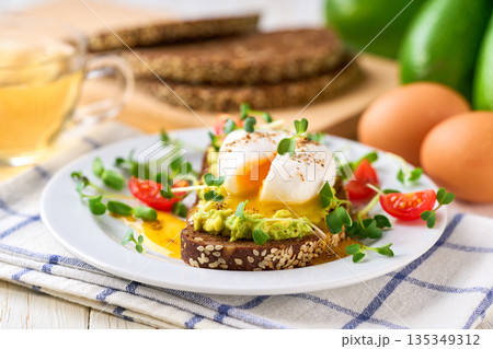Toast with avocado and egg with cup of tea for healthy breakfast or snack. Sandwich with avocado and poached egg on a white table, selective focus. Toast with avocado and egg with cup of tea for healthy breakfast or snack. Sandwich with avocado and poached egg on a white table, selective focus. 135349312