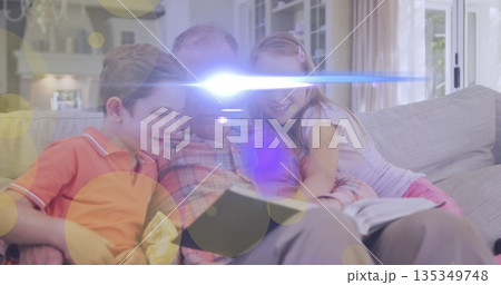 Reading grandfather sharing open book with grandchildren on sofa in living room, with fireplace 135349748