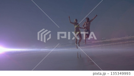 Running and leaping couple wearing swimsuit and trunks on wet sand with lens flare, ocean waves Running and leaping couple wearing swimsuit and trunks on wet sand with lens flare, ocean waves 135349993