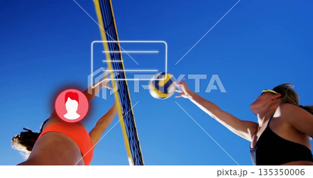 Jumping female volleyball players wearing sports tops hitting volleyball over net on beach court Jumping female volleyball players wearing sports tops hitting volleyball over net on beach court 135350006