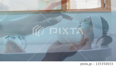 Soaking woman with green mask in white bathtub by window, esthetician dipping pad into cream jar Soaking woman with green mask in white bathtub by window, esthetician dipping pad into cream jar 135350033