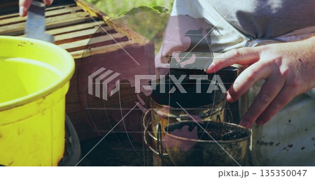 Operating beekeeper in grey zip-up top using bee smoker and hive tool at apiary with bucket 135350047