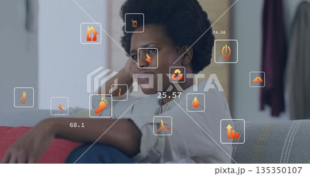 Sitting mid adult woman leaning on sofa arm in living room, with floating energy usage icons 135350107