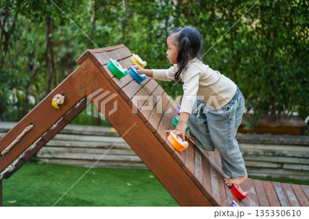 child girl practicing climbing on a wooden bouldering wall at outdoor playground child girl practicing climbing on a wooden bouldering wall at outdoor playground 135350610