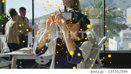 Seated woman wearing VR headset interacting with digital interface in office, with tablet, notebook 135352894