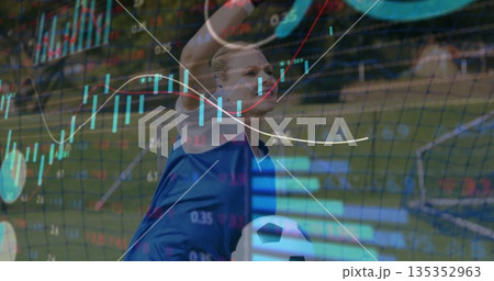 Raising arms goalkeeper in blue jersey catching ball on turf, financial charts overlaying net Raising arms goalkeeper in blue jersey catching ball on turf, financial charts overlaying net 135352963