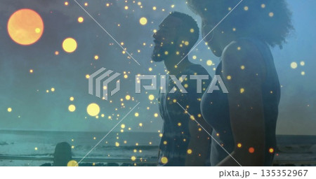 Gazing couple standing in swimwear on shoreline at dusk, with golden orbs, sun disc, wood posts 135352967