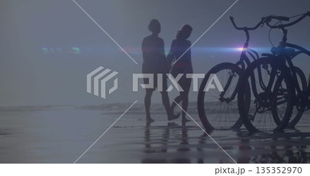 Walking couple holding hands in shallow water at shore, with cruiser bicycles reflecting on sand Walking couple holding hands in shallow water at shore, with cruiser bicycles reflecting on sand 135352970
