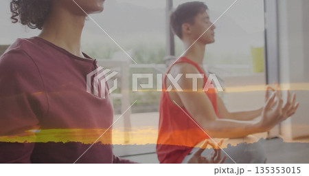 Friends wearing yogawear meditating crosslegged on mats in sunrise studio, with windows and plants 135353015