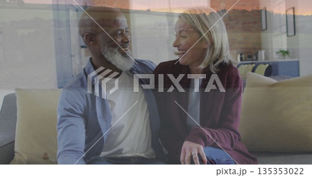 Smiling senior couple sitting closely on sofa in living room, with large window sunset view 135353022