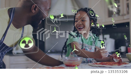 Father and daughter wearing aprons spreading sauce on pizza dough in kitchen, with eco icons 135353106