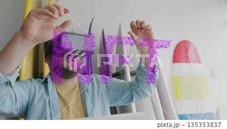 Man wearing VR headset raising hands interacting at surf shop with surfboards and laptop 135353837