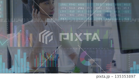Working agent wearing headset with microphone reviewing financial charts at office desk with laptop Working agent wearing headset with microphone reviewing financial charts at office desk with laptop 135353897