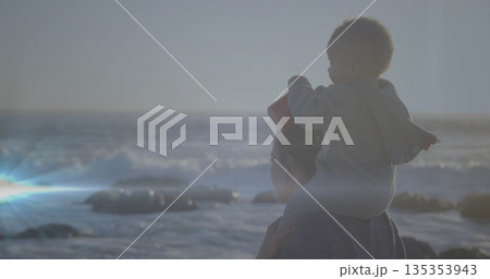 Standing mother carrying infant on shoulders by rocky shoreline at sunset, with red hat, copy space 135353943