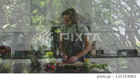 Adult man wearing striped apron slicing celery on kitchen island, with cutting board and tablet 135354065