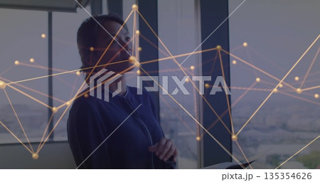 Standing woman wearing blue blazer near office window, holding tablet with digital network overlay 135354626
