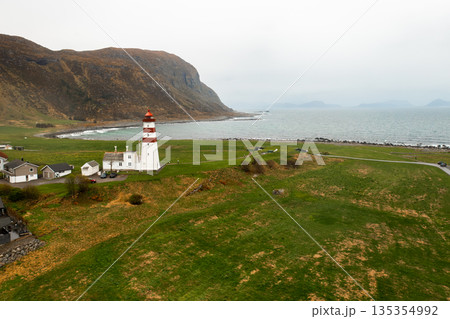 Nordic coastal scene with lighthouse, green meadow, sea, and distant mountains. 135354992