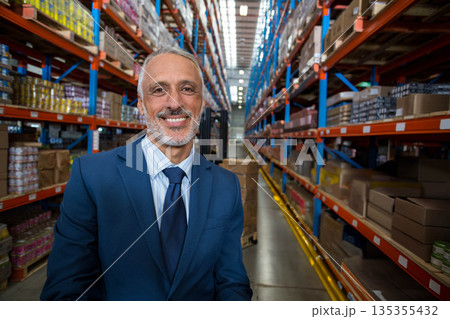 Senior man standing in warehouse aisle wearing suit and tie among shelving racks, boxes, copy space Senior man standing in warehouse aisle wearing suit and tie among shelving racks, boxes, copy space 135355432
