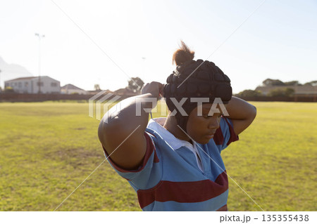 African American teenage girl wearing rugby jersey adjusting padded headguard on grass field African American teenage girl wearing rugby jersey adjusting padded headguard on grass field 135355438