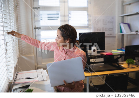 African American woman adjusting blinds at design studio holding laptop and clipboard, copy space African American woman adjusting blinds at design studio holding laptop and clipboard, copy space 135355466
