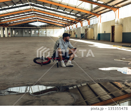 Man sitting astride red and black BMX bike holding smartphone inside vacant warehouse with puddles 135355478