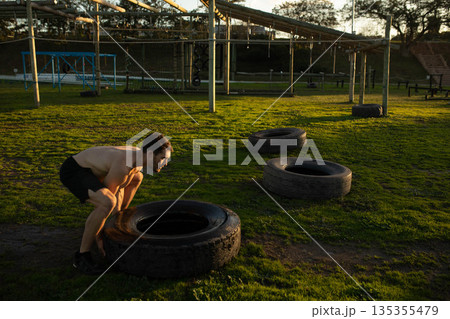 Man wearing black shorts squatting on field flipping tractor tire near obstacle frame, copy space 135355479