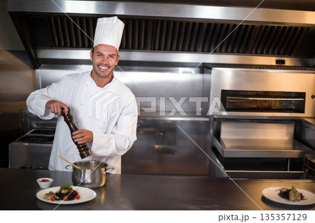 Male chef seasoning with wooden pepper mill over saucepan on steel counter in commercial kitchen 135357129