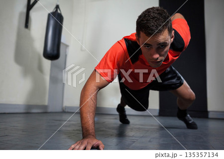 Male in red shirt performing one-arm pushup on rubber gym floor with punching bag, wall pad 135357134