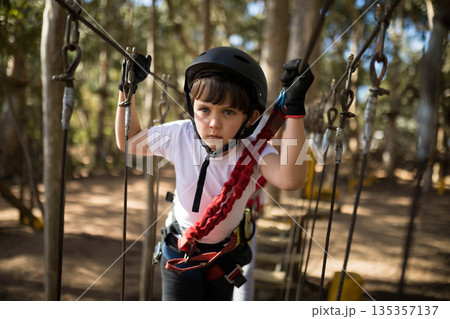 Boy navigating suspended rope course in woods wearing black helmet, gloves, red black harness 135357137