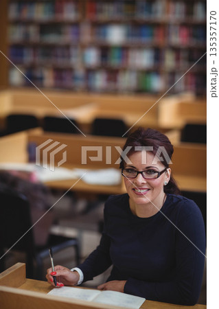Mid adult woman sitting at library carrel wearing eyeglasses holding pen over open book, copy space 135357170