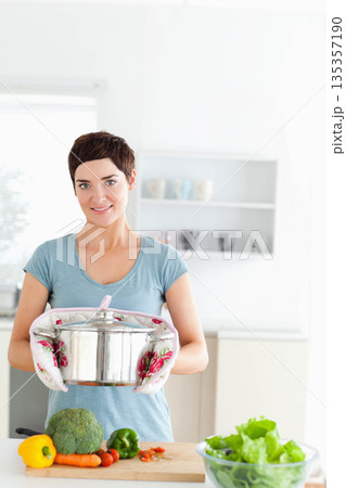 Woman holding large steel pot with floral oven mitts in home kitchen with broccoli peppers carrots 135357190