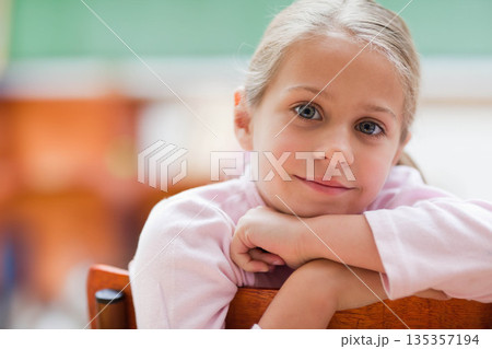 Female child wearing pink top leaning arms on wooden chair and gazing in classroom with chalkboard Female child wearing pink top leaning arms on wooden chair and gazing in classroom with chalkboard 135357194