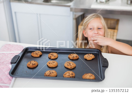 Blonde-haired female child standing at kitchen counter reaching for cookie on tray, copy space Blonde-haired female child standing at kitchen counter reaching for cookie on tray, copy space 135357212