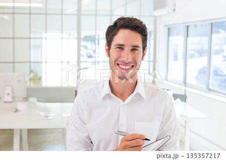 Man wearing white shirt standing at center of office holding notebook and pen, featuring monitors Man wearing white shirt standing at center of office holding notebook and pen, featuring monitors 135357217