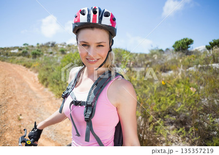 Smiling woman wearing cycling helmet, gloves, backpack standing on dirt trail holding handlebars Smiling woman wearing cycling helmet, gloves, backpack standing on dirt trail holding handlebars 135357219
