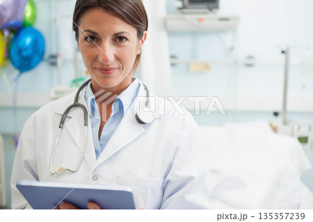 Female physician standing in hospital holding clipboard wearing stethoscope near balloons by bed Female physician standing in hospital holding clipboard wearing stethoscope near balloons by bed 135357239