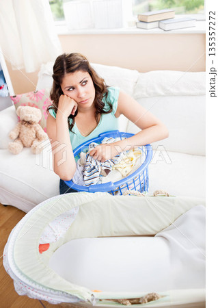 Woman sitting on white sofa holding blue laundry basket with baby clothes near bassinet 135357272