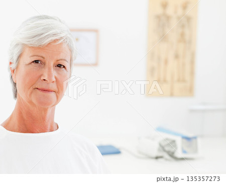 Senior woman standing in exam room with skeleton poster, certificate and BP monitor, copy space 135357273